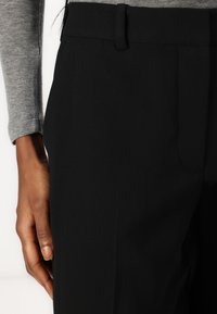 Pantalon noir avec une texture lisse, présentant un devant plat, des passants pour ceinture et une poche latérale. Vue rapprochée avec un haut à manches longues gris.