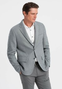 Grau gestreifter Blazer mit Revers, zwei vorderen Taschen und taillierter Passform, getragen über einem weißen Hemd mit schwarzen Knöpfen und grauen Hosen.