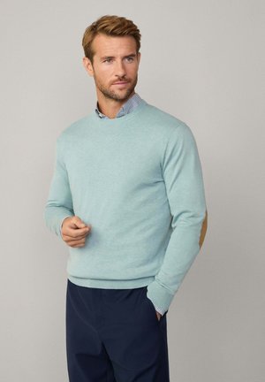 Helles blaues Strickpullover mit gerippten Saum, ausgestattet mit braunen Ellbogenpatches. Über einem blau-weißen karierten Hemd getragen; das Modell befindet sich in einer neutralen Umgebung.