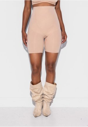 Frau trägt hoch taillierte, hautfarbene Shorts und lässige beige Stiefel und steht vor einem schlichten weißen Hintergrund.