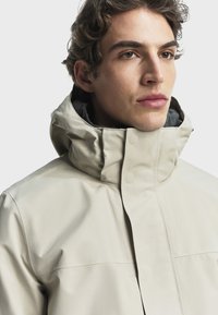 Bež jakna otporna na vodu s visokim ovratnikom, koja se odlikuje glatkom teksturom, kopčama na dugme i elegantnim, modernim dizajnom. Siva podstava vidljiva.