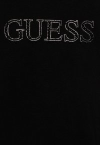 Mustapaita, jossa on sana "GUESS" koristeltuna hopea- ja mustilla strasseilla, keskitettynä sileän kankaan tekstuuria vasten.