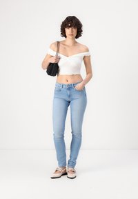 Bela off-shoulder majica s valovitim robom, svetlo modre skinny traperice in bež balerinke s trakovi. Model drži majhno črno torbico.