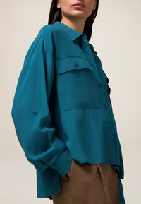 Cămașă teal cu butoni dintr-un material ușor, cu o croială relaxată, având două buzunare la piept și mâneci suflate. Complementată de pantaloni maro.