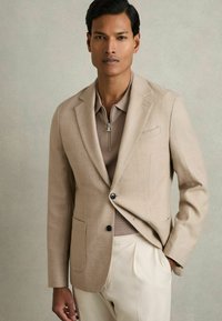 Sacou texturat, de culoare bej deschis, cu două buzunare frontale, un singur nasture negru și rever ciupit, purtat peste un tricou polo de culoare taupe.