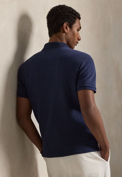 Νέος άνδρας που στέκεται με τα χέρια στις τσέπες, φορώντας μπλε σκούρο πόλο με κοντά μανίκια και ανοιχτόχρωμο παντελόνι μπροστά σε ουδέτερο τοίχο.