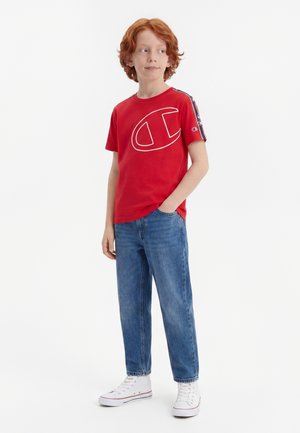 Garçon roux portant un t-shirt rouge à manches courtes avec un logo, un jean bleu et des baskets blanches montantes, debout, une main dans la poche, regardant sur le côté.