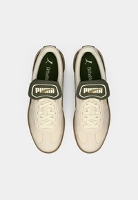 Sapatilhas de cor creme com um upper texturizado, atacadores e uma faixa de Velcro verde com um logo dourado. Sola de borracha com palmilha Ortholite.