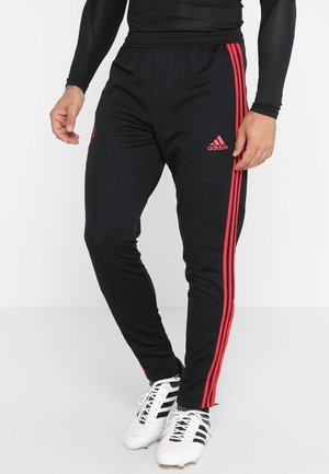 Homme portant un pantalon de survêtement Adidas noir avec des bandes rouges sur les côtés et des chaussures à crampons blanches Adidas, debout devant un fond uni.