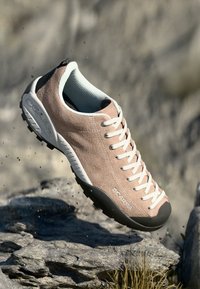 Helle rosa Wildlederschuhe mit einem hellblauen Stoffkragen, weißen Schnürsenkeln und einer schwarzen Gummisohle. Über steinigen Untergrund erhoben. Markenname sichtbar.