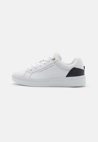 Zapatillas blancas de caña baja fabricadas en material sintético suave, con un acento negro en el talón y un detalle de logo dorado en el costado. Suela blanca texturizada.