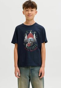 Garçon portant un t-shirt de Noël bleu marine avec un crâne barbu en chapeau rouge tenant une tasse, associé à un jean bleu ample.