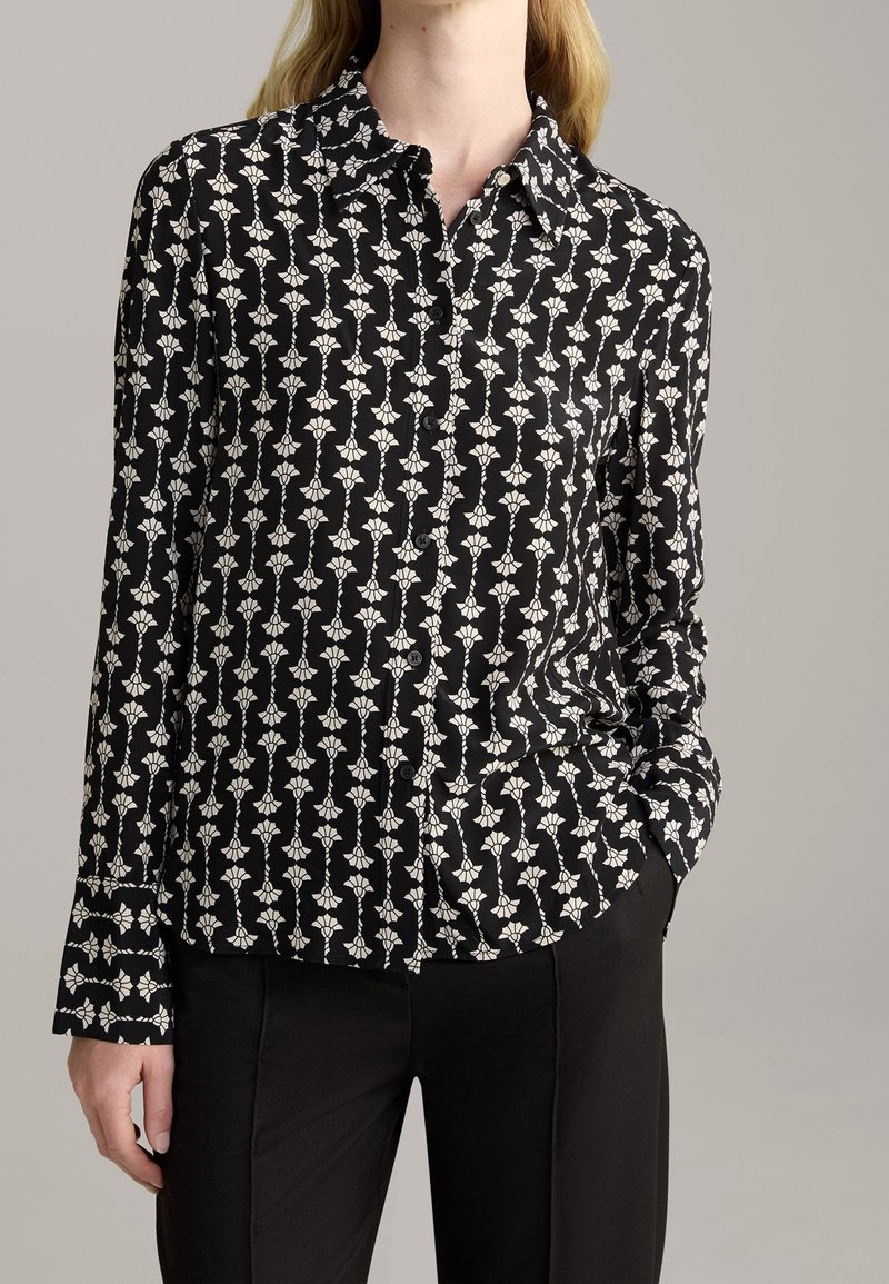 Black button-up shirt with a white floral pattern, long sleeves, and a collar. Smooth fabric with a fitted design. Paired with black pants.