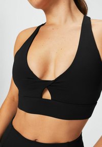 Zwarte sportbeha met een diepe V-hals en een kruisdetail aan de voorkant, gemaakt van een soepele stof. Heeft een uitsnijding onder de buste en brede banden.