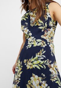 Marineblauwe bloemenjurk met gele en witte bloesems, aansluitende lijfje met plooien en een vloeiende rok. Gemaakt van een lichte stof.