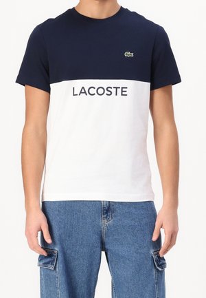 Person iført en marineblå og hvid Lacoste t-shirt med logo og tekst foran, kombineret med blå denim cargobukser.