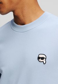 Lichtblauwe sweatshirt met een geribbelde ronde halslijn; heeft een zwart-wit rubberen patch van een gezicht met een zonnebril op de borst.