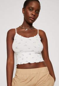 Biele rebrované crop top s vlnitým lemovaním a modrou kvetinovou potlačou. Tenké ramienka a priliehavý strih. Kombinovaný s béžovými šortkami na elastický pás.