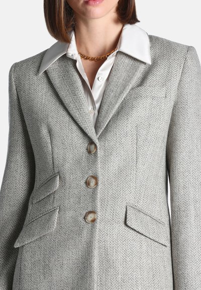 Femme portant un blazer gris à chevrons avec trois boutons, des poches à rabat, une chemise blanche colée et un collier chaîne dorée.
