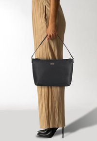 Geantă de umăr din piele texturată neagră, cu un mânere scurt, de formă rectangulară și un logo subtil. Asortată cu pantaloni plisati bej și tocuri negre.