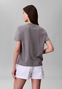 Hosszú sötét hajú nő egyszínű szürke rövid ujjú pólóban és fehér felhajtott szegélyű rövidnadrágban, háttal áll egy szürke háttér előtt.