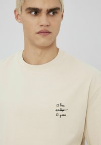 T-shirt en coton beige avec un graphique comprenant des cases à cocher et du texte. Design simple à col rond, manches courtes et texture lisse.