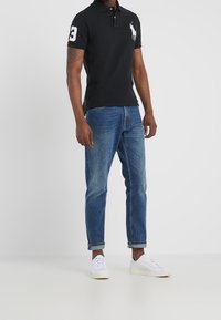 Man med svart polo med vitt logo, blå uppkavlade jeans och vita sneakers som står mot en enfärgad bakgrund.