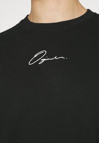 Camiseta de algodón negra con cuello redondo y dobladillo acanalado, que presenta un diseño de firma manuscrita blanca en la zona del pecho.