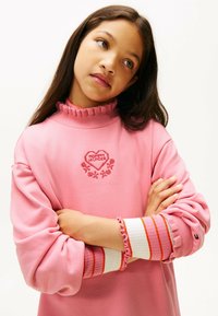 Sudadera rosa con cuello con volantes, "MAMÁ" bordado en un corazón rojo, detalles florales y acentos a rayas en los puños con una textura suave.