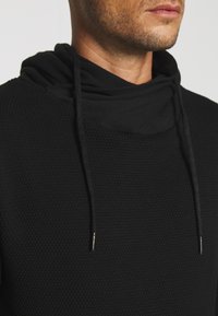 Sudadera negra con una tela texturizada, que cuenta con un cuello alto y cordones con puntas metálicas. Vista de cerca del área del cuello y el hombro.