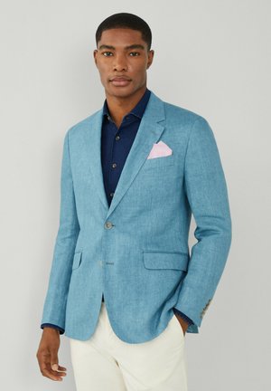 Blazer en lin bleu clair avec un col cranté, deux poches avant et un mouchoir de poche rose. Porté par-dessus une chemise bleu foncé.