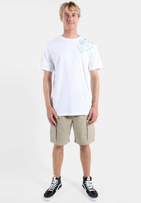 T-shirt blanc en coton avec un motif floral bleu sur le côté supérieur gauche, associé à un short kaki et des baskets montantes noires.