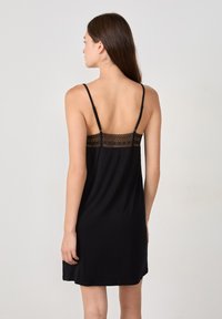 Donna con lunghi capelli castani che sta in posizione di spalle, indossando un abito senza maniche nero con dettagli in pizzo e sottili spalline.