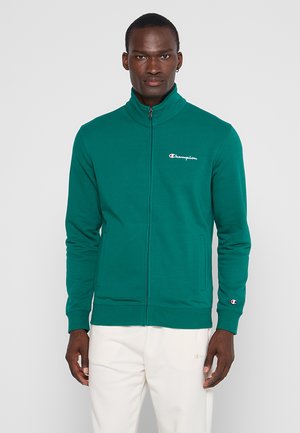 Uomo che indossa giacca verde Champion con zip e pantaloni bianchi, in piedi davanti a uno sfondo grigio chiaro uniforme.