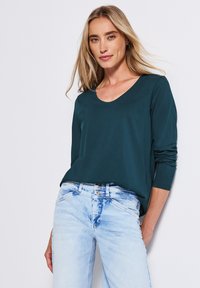 Teal lange mouwen top met een ronde halslijn, gecombineerd met lichtblauwe gescheurde jeans. Soepele textuur, casual pasvorm en minimale details.