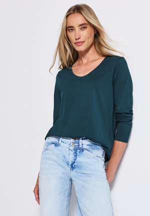 Maglietta a maniche lunghe color teal con scollo a barca, abbinata a jeans chiari strappati. Texture liscia, vestibilità casual e dettagli minimi.
