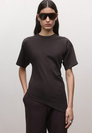 Kvinde iført overdimensionerede mørke solbriller, asymmetrisk mørkebrun kortærmet top og matchende bukser, stående mod en ensfarvet baggrund.