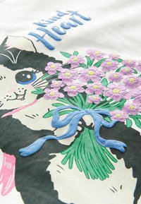 Grafikus póló, amelyen egy fekete-fehér macska látható, amint egy lila virágcsokrot tart, kék szalaggal, és az "Aranyos Szív" felirat olvasható.