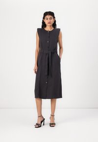 Vestido sem mangas cinza escuro com cintura cingida por cinto, frente com botões e ombros estruturados. Combinado com sandálias pretas de salto alto decoradas com laços.