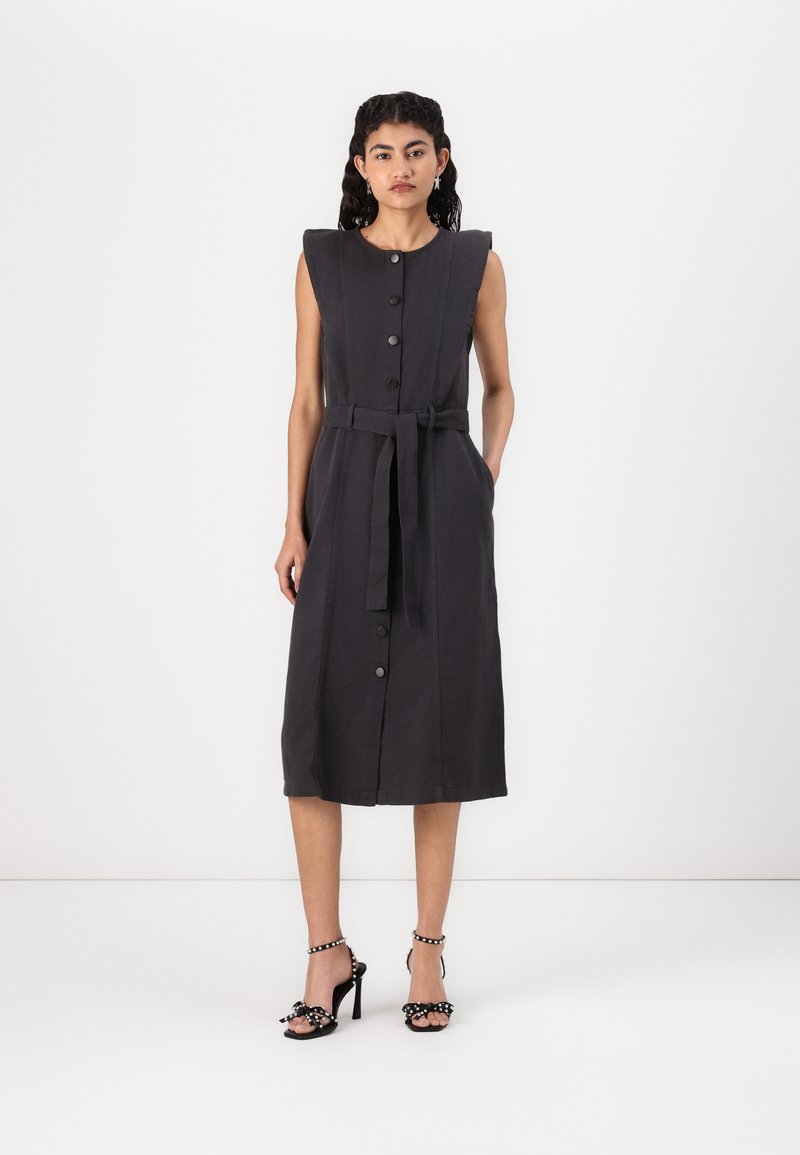 Dunkelgraues ärmelloses Kleid mit tailliertem Schnitt, Knopfleiste und strukturierten Schultern. Kombiniert mit schwarzen Absatzsandalen mit Schleifen.