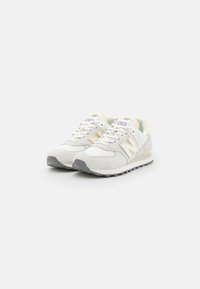 Chaussures de sport en daim blanc et mesh avec des accents jaunes, dotées d'une semelle texturée, d'un bout arrondi et du logo « N » sur le côté.