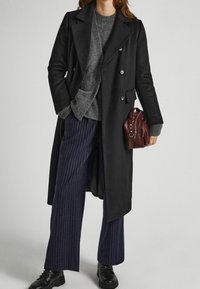 Abrigo negro de doble botonadura con botones azules, suéter gris de punto, pantalones azul marino de rayas finas y un pequeño bolso color burdeos. Botas negras con cordones.