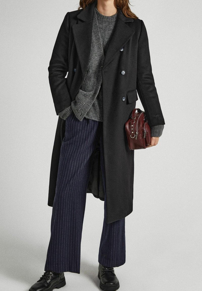 Abrigo negro de doble botonadura con botones azules, suéter gris de punto, pantalones azul marino de rayas finas y un pequeño bolso color burdeos. Botas negras con cordones.