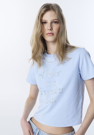 Giovane donna con lunghi capelli biondi che indossa una maglietta corta di colore azzurro chiaro con la scritta "Under the Familiar Sun" composta da puntini multicolori.