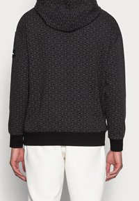 Sudadera negra con un patrón de logo repetido en toda la prenda, con un bolsillo tipo canguro y puños de canalé. La tela es suave y de textura lisa.