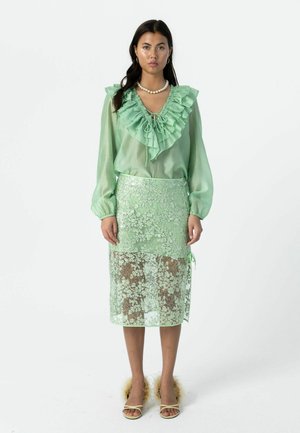 Naine, kes kannab mintrohelise värvi volangidega pluusi ja sobivat pitsilise seelikut, lisaks pärlikaelakeed ja läikivate varbavahekingadega jalanõusid.