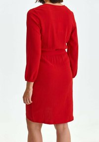 Robe rouge texturée à manches longues, avec une taille cintrée et une coupe mi-longue, dotée d'un col rond et de fentes latérales discrètes.