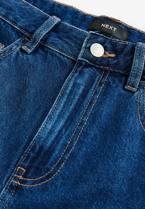Tumed sinine denimist teksapüksid siledäbise tekstuuriga, millel on eesneveti, nööbi kinnitamine ja oranžad õmblused. Silti peal on kirjas "NEXT."