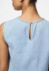 Světle modrý denimový top s kulatým výstřihem, bez rukávů a s klíčovou dírkou na zádech, která je zajištěna modrým knoflíkem.