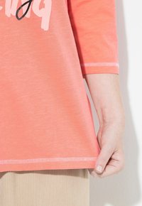 Person in einem korallpinken Langarmshirt mit hellrosa und schwarzem Text, beigen, gerippten Hosen, der den Saum des Shirts in der Hüfthöhe hält.